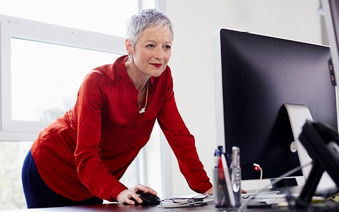Woman leaning over desk holding mouse looking at computer monitor.