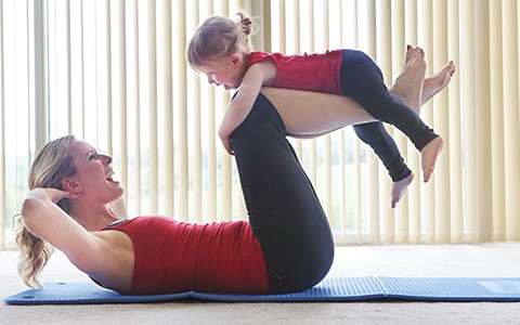 Woman lying down on a yoga mat with a child holding onto her legs.